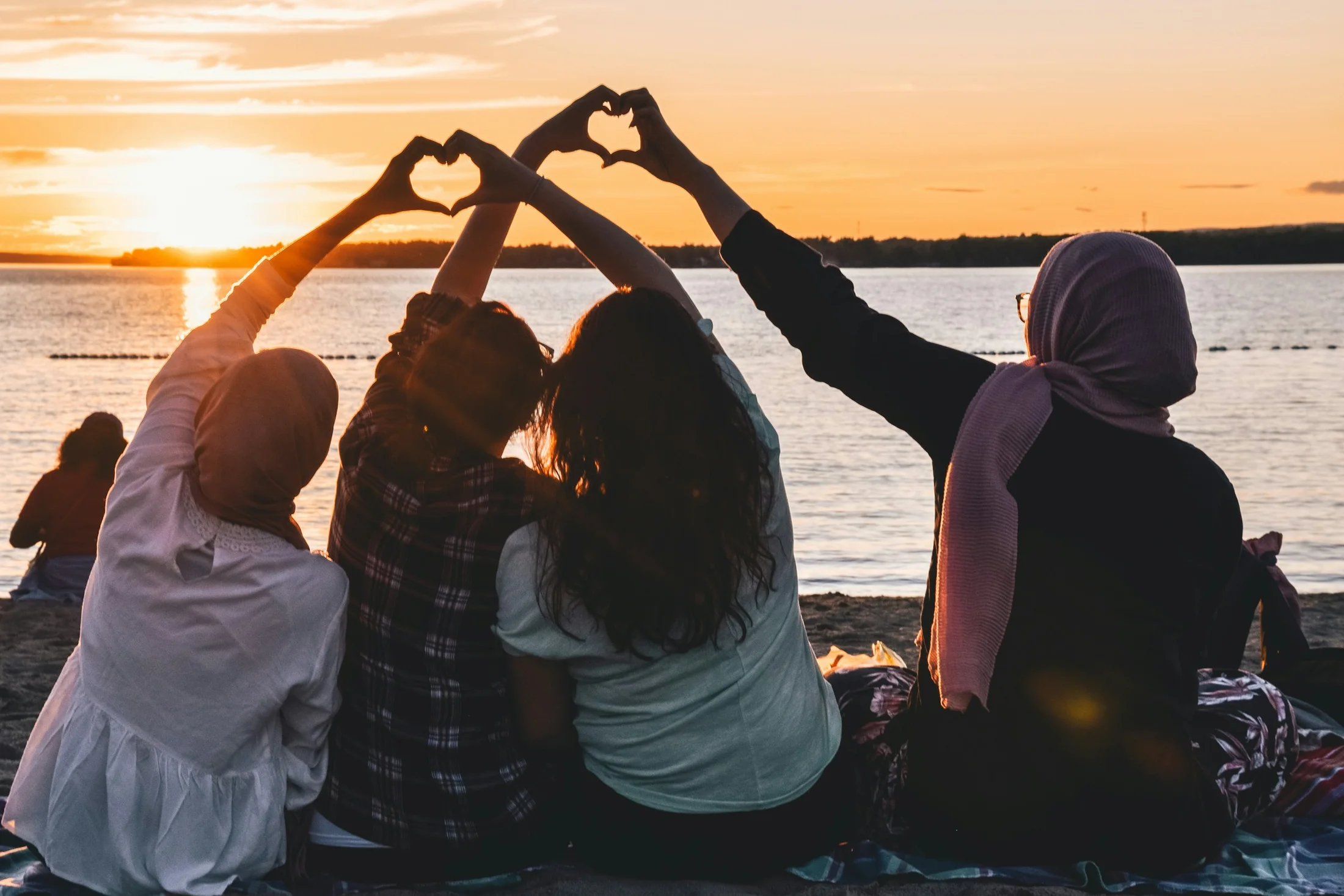 Four friends sitting by the sea at sunset, making heart shapes with their hands